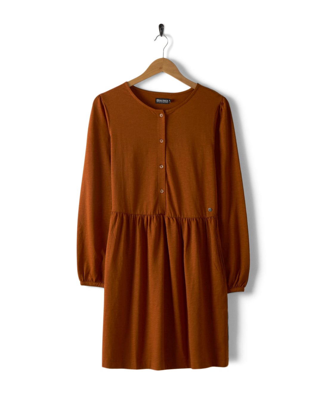The Winnie women's dress in dark orange from Saltrock, crafted from Jersey slub fabric, features long sleeves, buttons down the front, and an elasticated waist, elegantly displayed on a wooden hanger against a crisp white background.