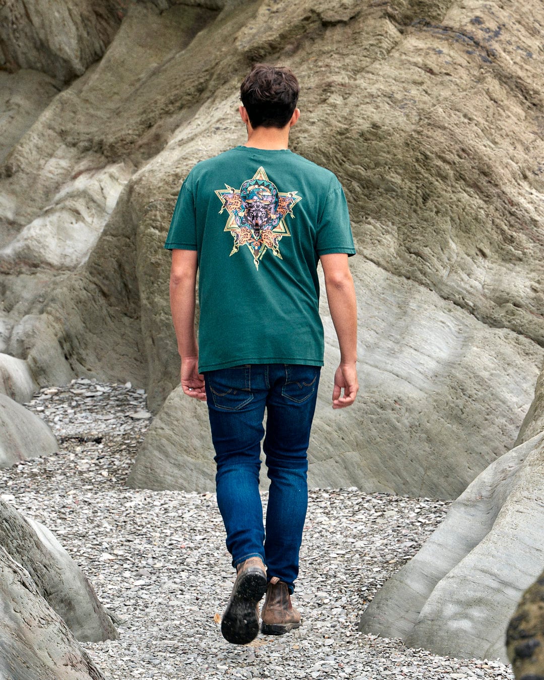 A man walking down a rocky path wearing a Saltrock Wolfscale - Mens Short Sleeve T-Shirt - Dark Green with a snake graphic.