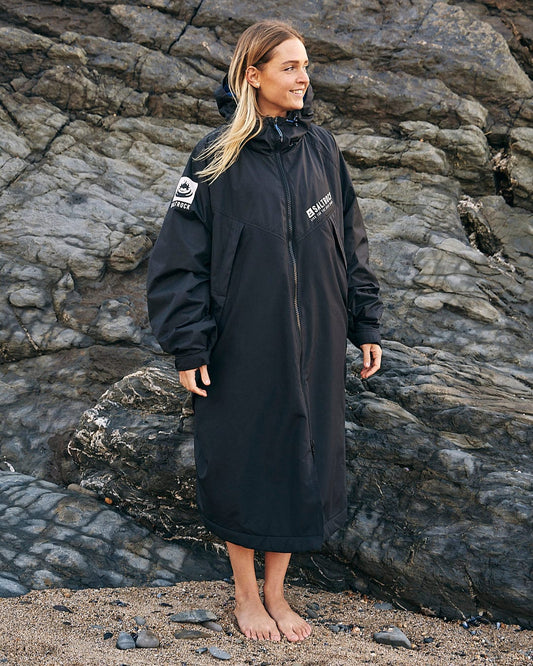 A woman in a Saltrock Four Seasons - Waterproof Changing Robe - Black/Blue standing on a rocky beach.