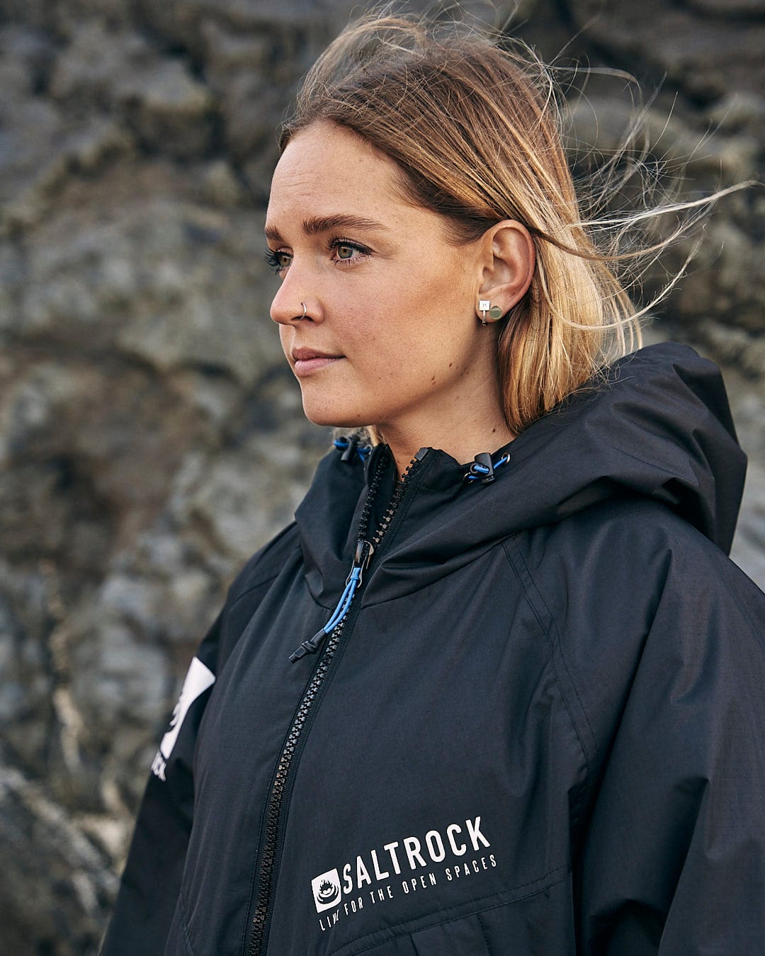 A high-performance woman in a Saltrock - Four Seasons Waterproof Changing Robe in Black/Blue standing in front of rocks.