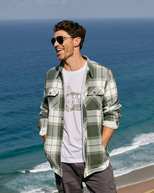 A man standing on a beach wearing sunglasses and a flannel shirt with Saltrock's Woody - Mens Borg Lined Shacket - Green Check branding.
