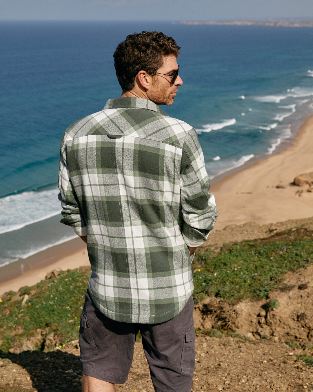 A man is standing on a hill overlooking the ocean, mesmerized by the vastness of the Saltrock Woody - Mens Borg Lined Shacket - Green Check.