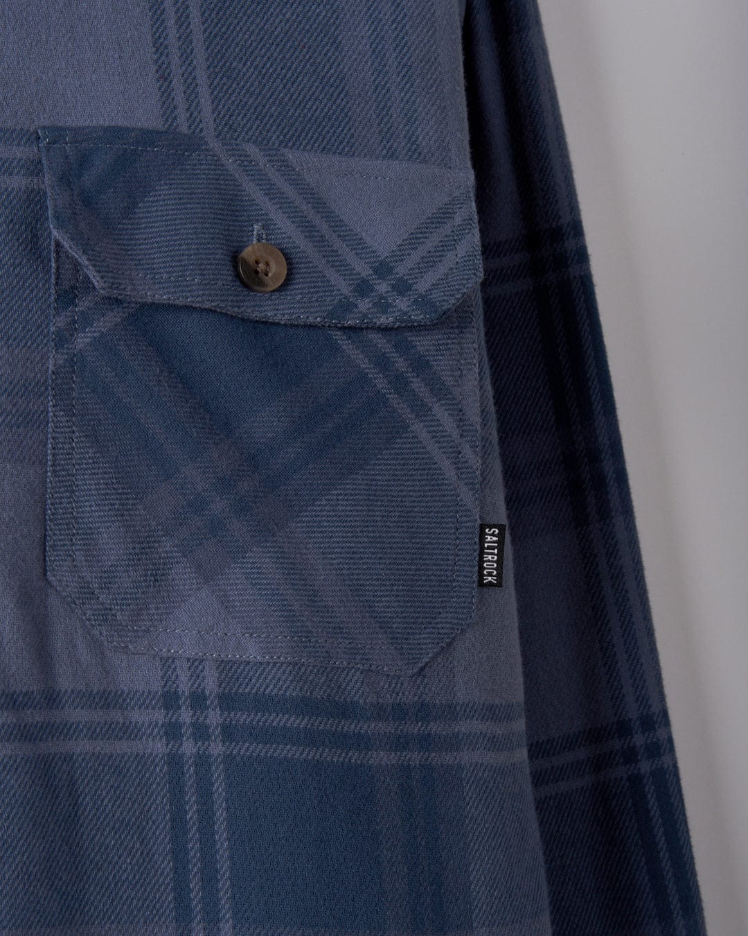 A close-up of the Woody Men's Sherpa Lined Shacket in Mid Blue by Saltrock showcases a button and a small black tag, stylishly crafted from recycled materials.