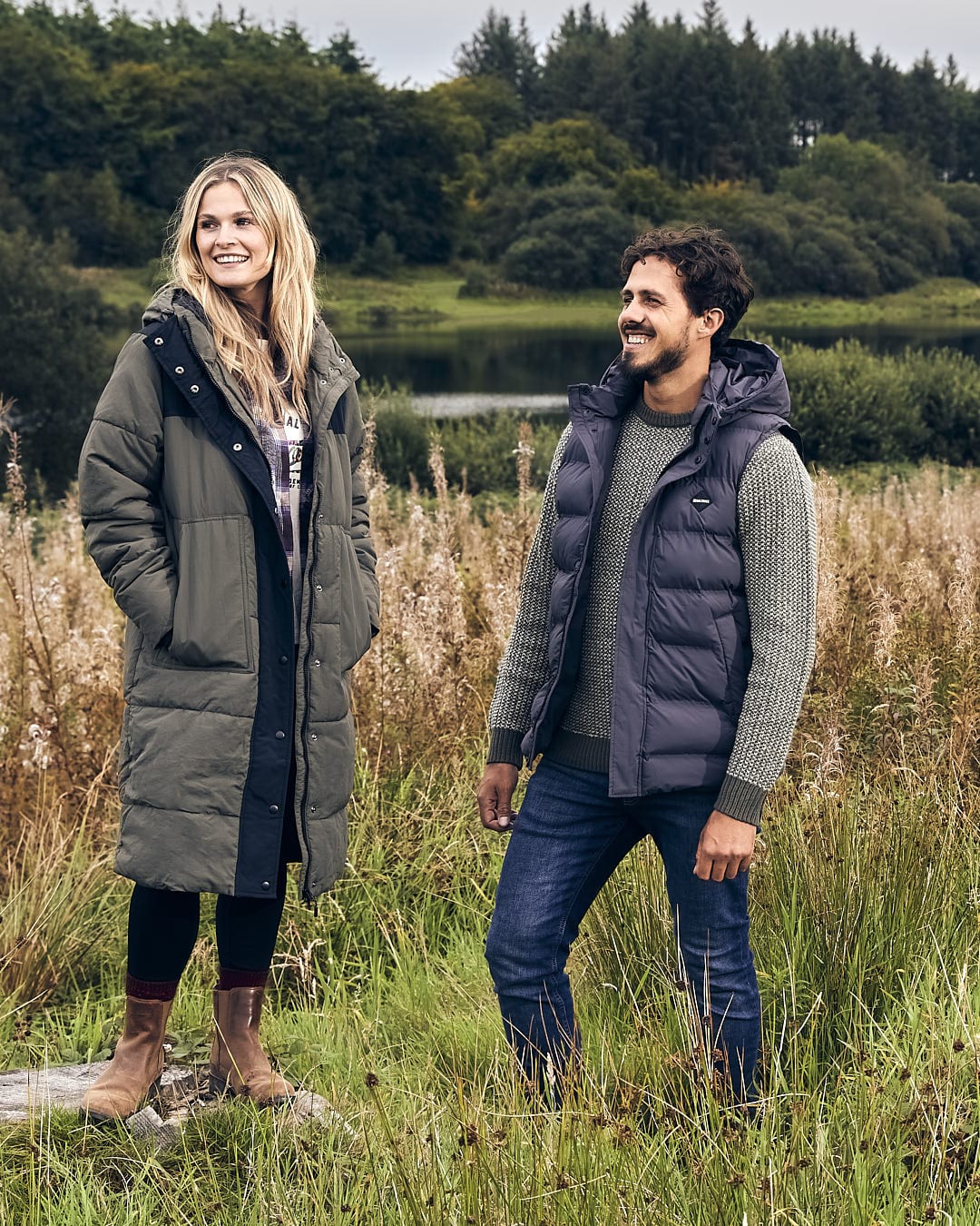 A man and woman, wearing Saltrock's Xavier - Mens Padded Gilet in Dark Grey, standing in a field near a lake, wearing machine washable clothes.