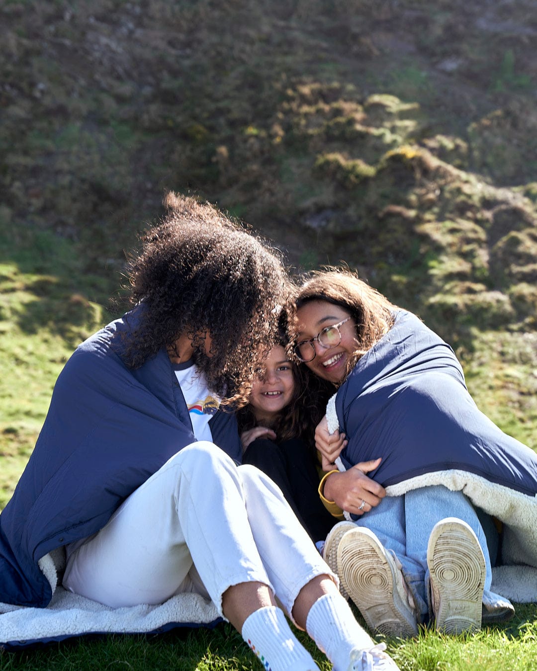 Three women wrapped up in Saltrock's Yogi - Camping Blanket - Blue on a grassy field.