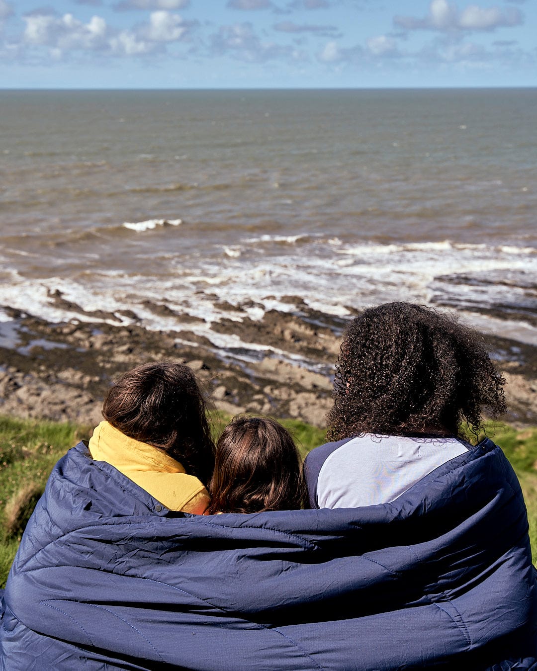 A group of people sitting on a Yogi - Camping Blanket - Blue by Saltrock looking at the ocean.