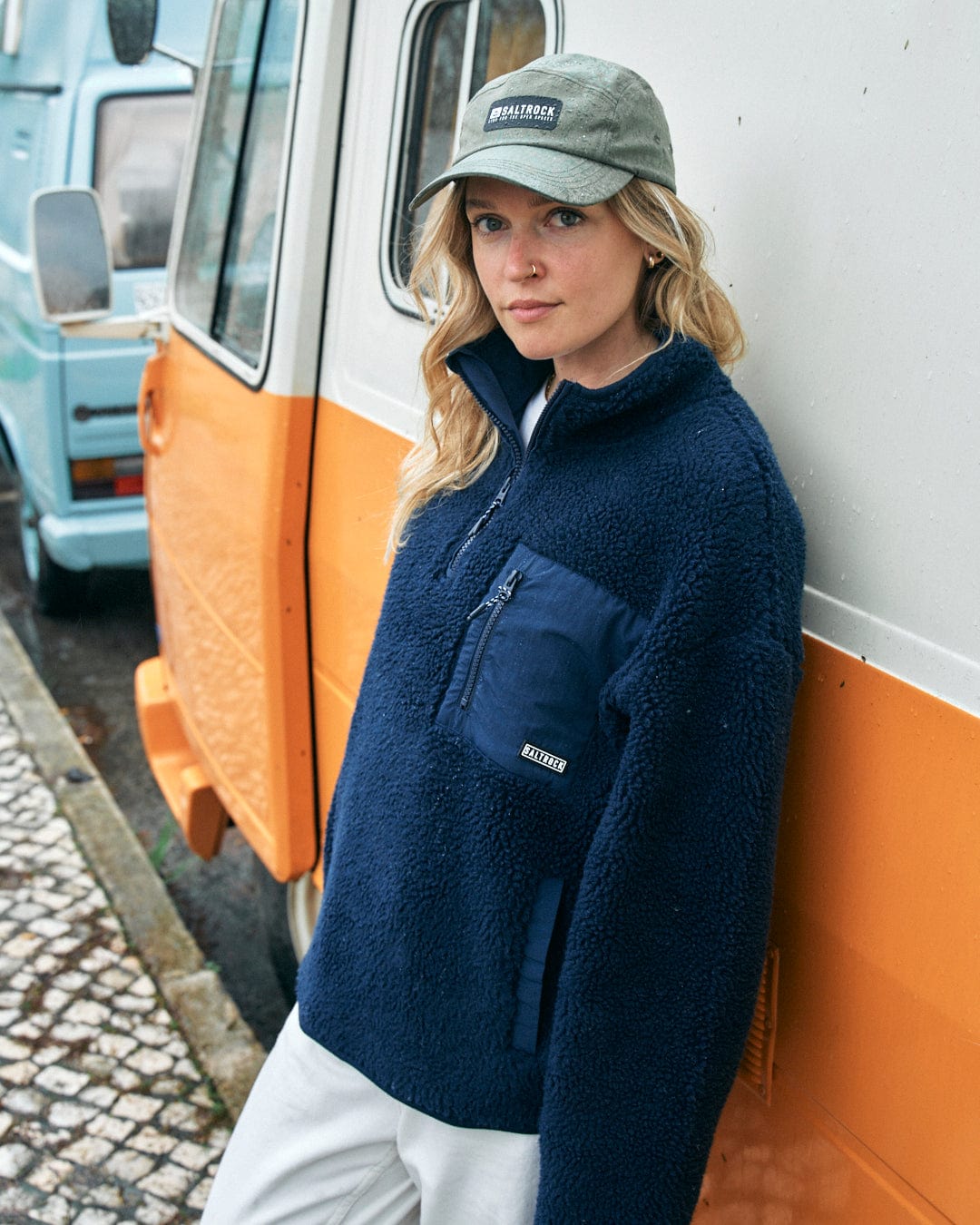 A woman wearing a blue fleece jacket and a Gaitor 5 Panel UPF cap by Saltrock stands in front of an orange and a blue van.