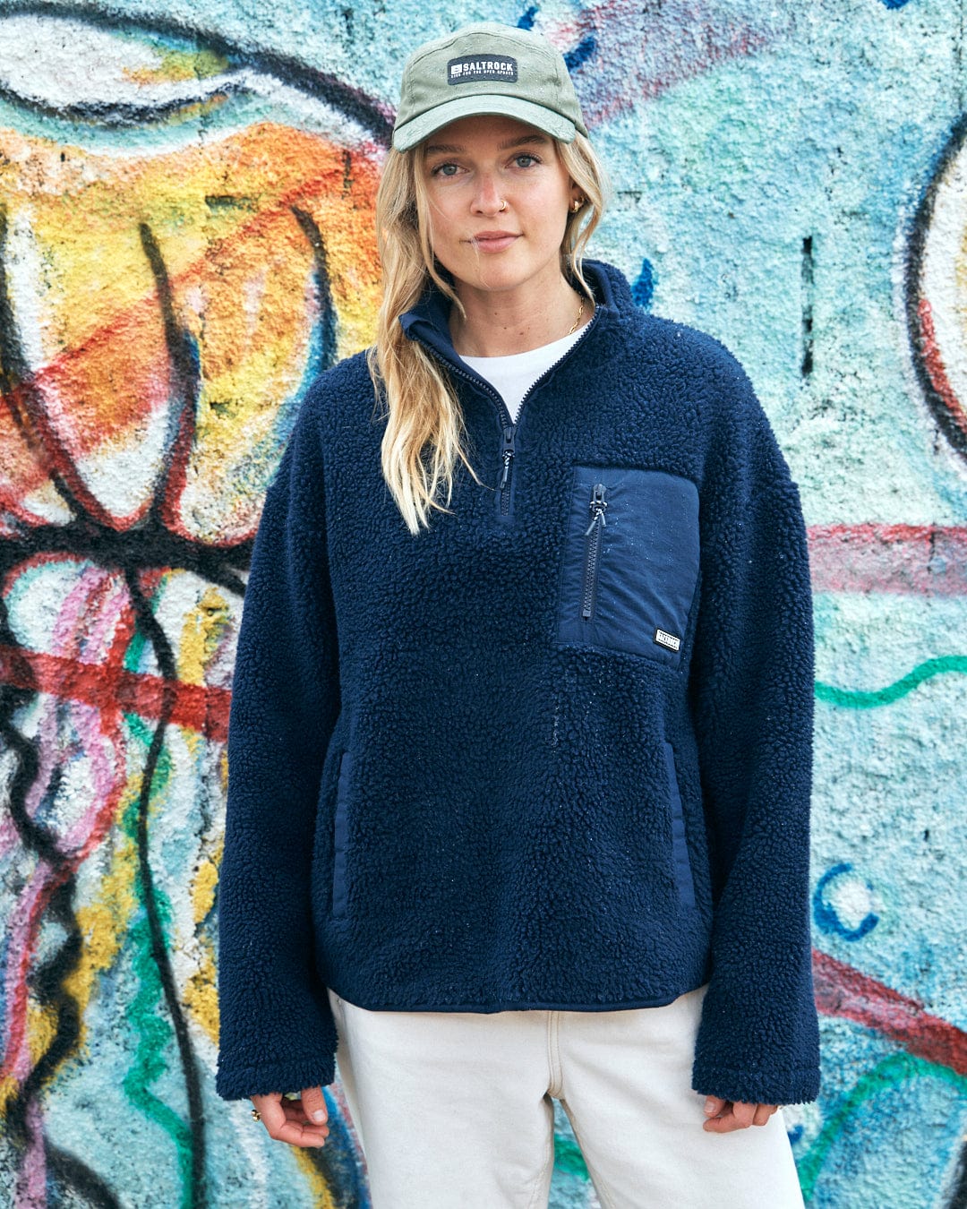 A woman wearing a Saltrock-branded fleece jacket and a Gaitor 5 Panel UPF Cap - Green stands in front of a graffiti-covered wall.