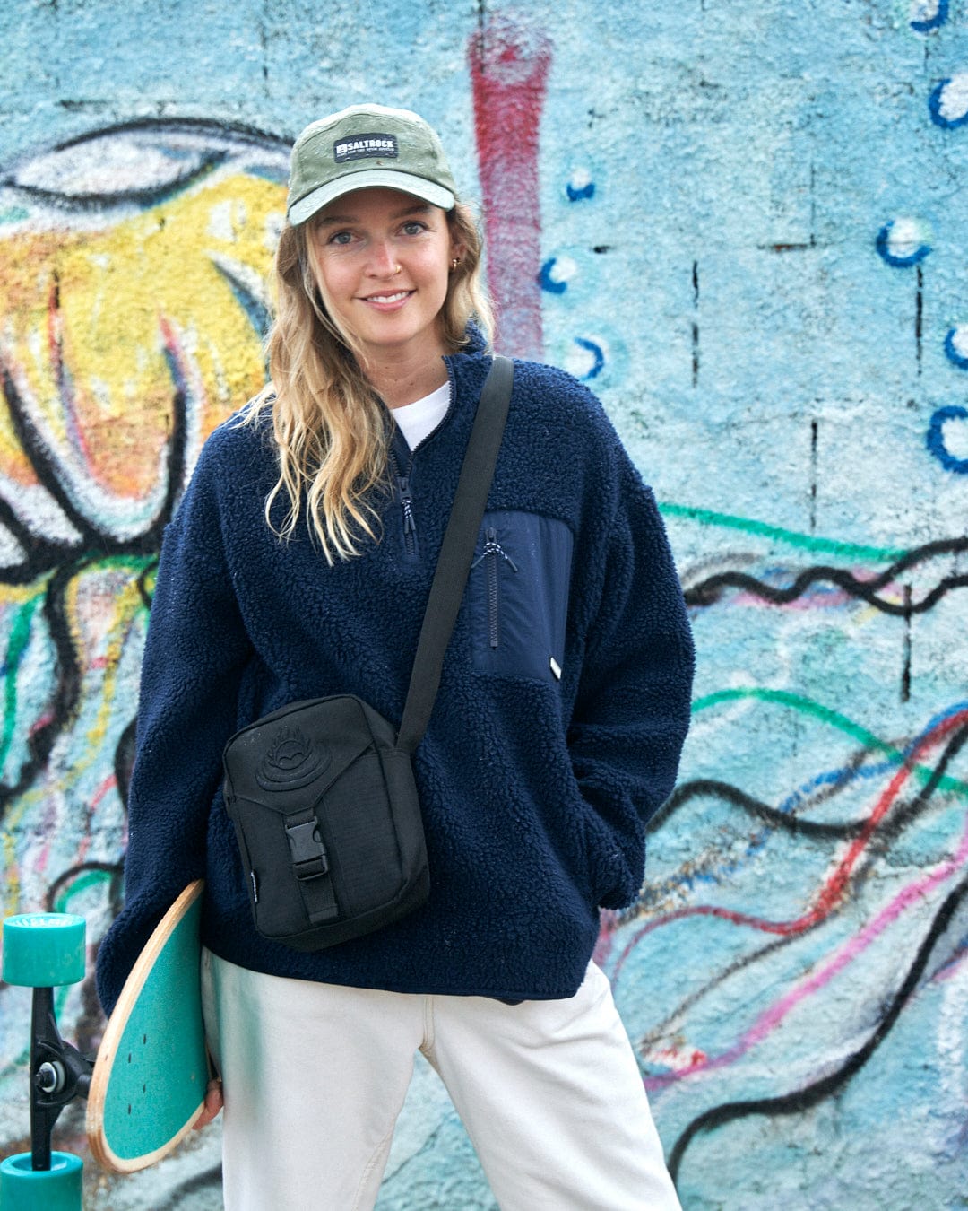 A woman holding a Zella - Womens 1/4 Neck Fleece - Dark Blue by Saltrock in front of a graffiti wall with zip details.