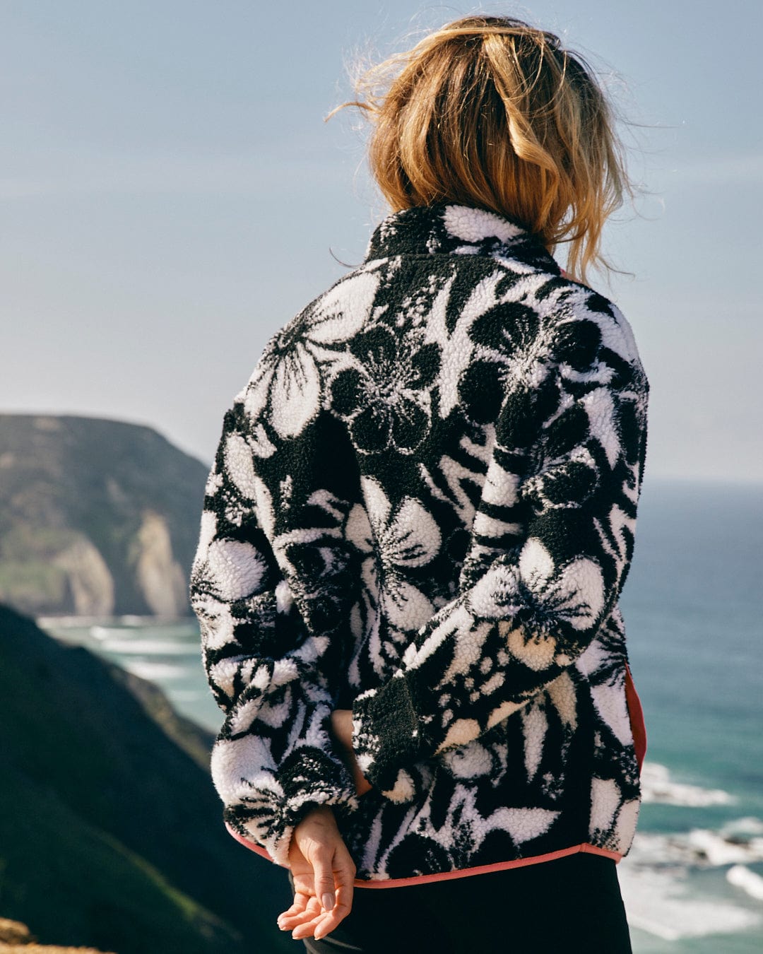 A woman wearing a Saltrock Zella Hibiscus - Womens 1/4 Neck Fleece - Black Floral jacket on a cliff overlooking the ocean.