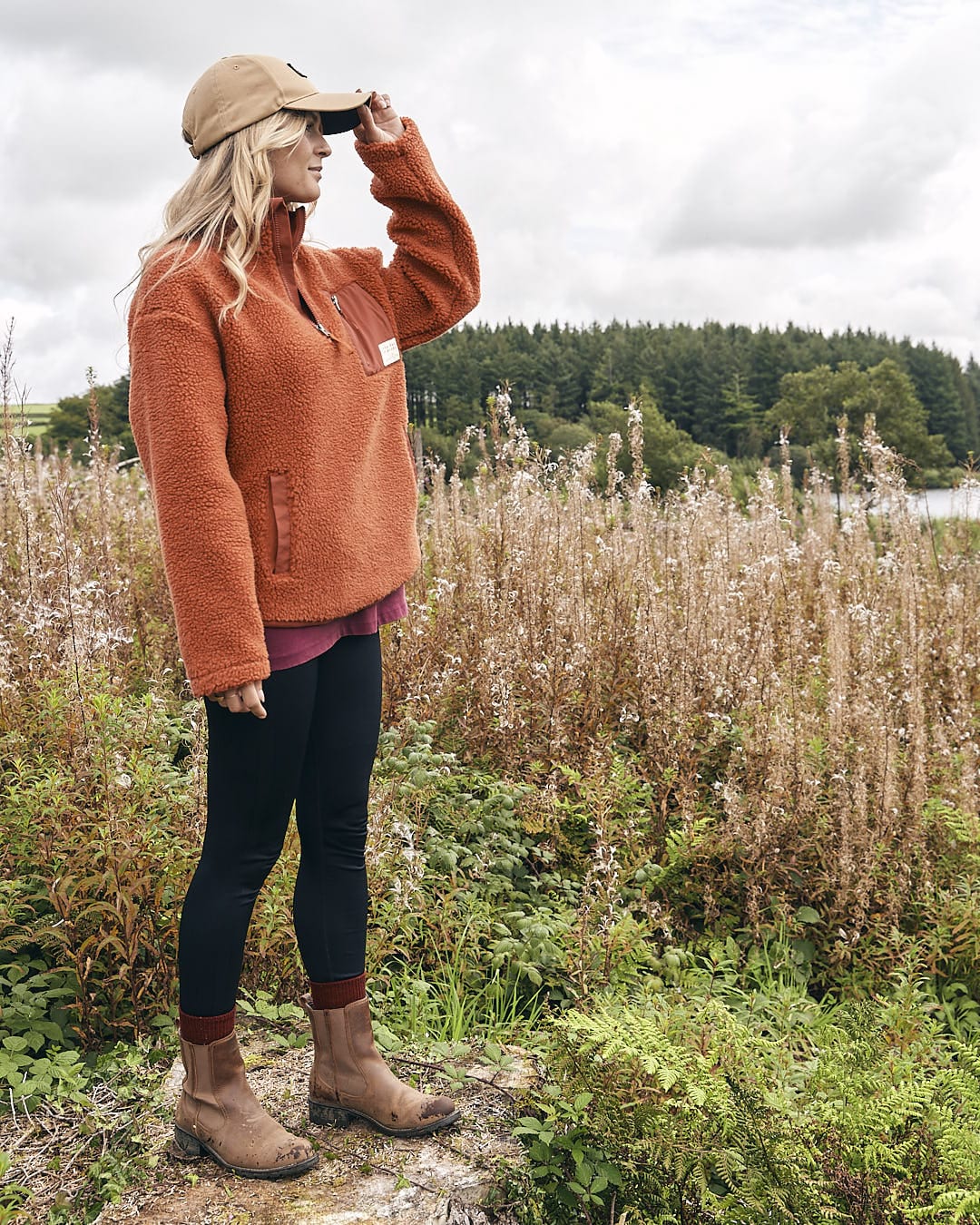 A woman standing in a field wearing a Saltrock Zella Solid - Womens Borg Fleece - Burnt Orange sweater and hat.