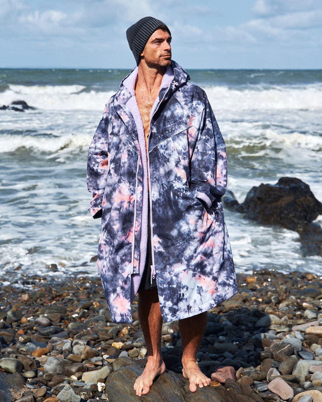 A man in a Saltrock Recycled Changing Robe - Pink Tie Dye and a beanie stands barefoot on the rocky beach, gazing into the distance. Waves crash under a cloudy sky, contrasting with his vibrant attire.