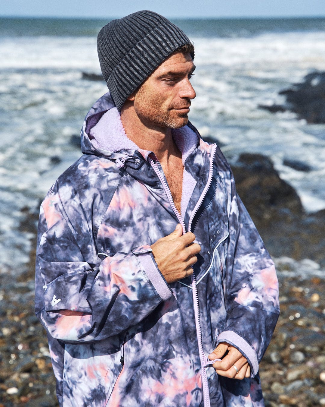 A person in a Saltrock Recycled Changing Robe in Pink Tie Dye and a gray beanie stands on a rocky beach with the ocean in the background.