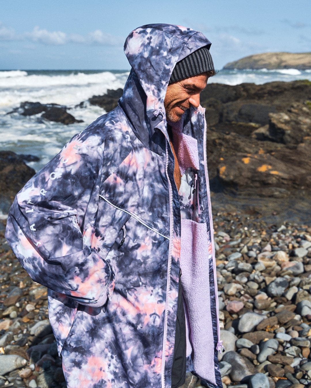 A person wearing the Saltrock Recycled Changing Robe in Pink Tie Dye and a beanie stands on a rocky beach with waves and hills in the background.