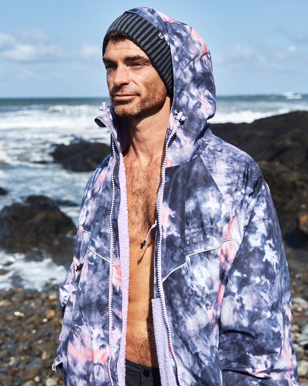 A person wearing a Saltrock Recycled Changing Robe in pink tie dye and a striped beanie stands on a rocky beach, gazing at the sea.
