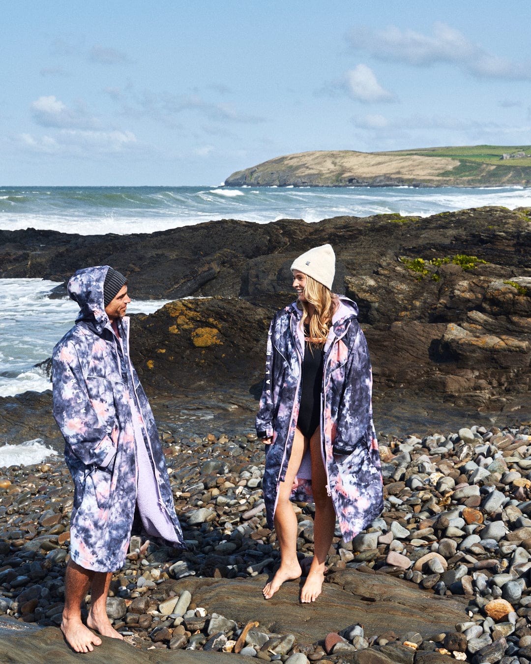 Two people stand on a rocky beach, wearing Saltrock's Recycled Changing Robe in Pink Tie Dye and beanies, with the ocean and a grassy hill as a serene backdrop.