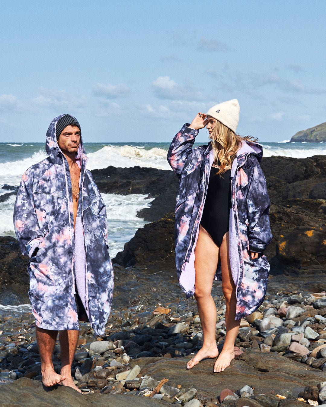 Two people stand on a rocky beach in Saltrock's Recycled Changing Robe - Pink Tie Dye. One wears a beanie and swimsuit, the other sports their hood up with shorts. The vibrant robes, made from recycled materials, blend seamlessly with the waves and cloudy sky behind them.