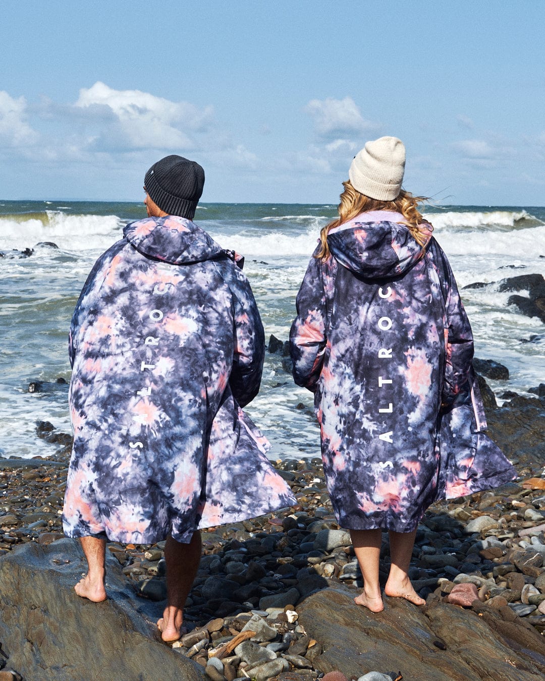 Two individuals in waterproof, pink tie-dye Saltrock recycled changing robes stand on the rocky beach facing ocean waves, with "ALTROS" visible on the back.