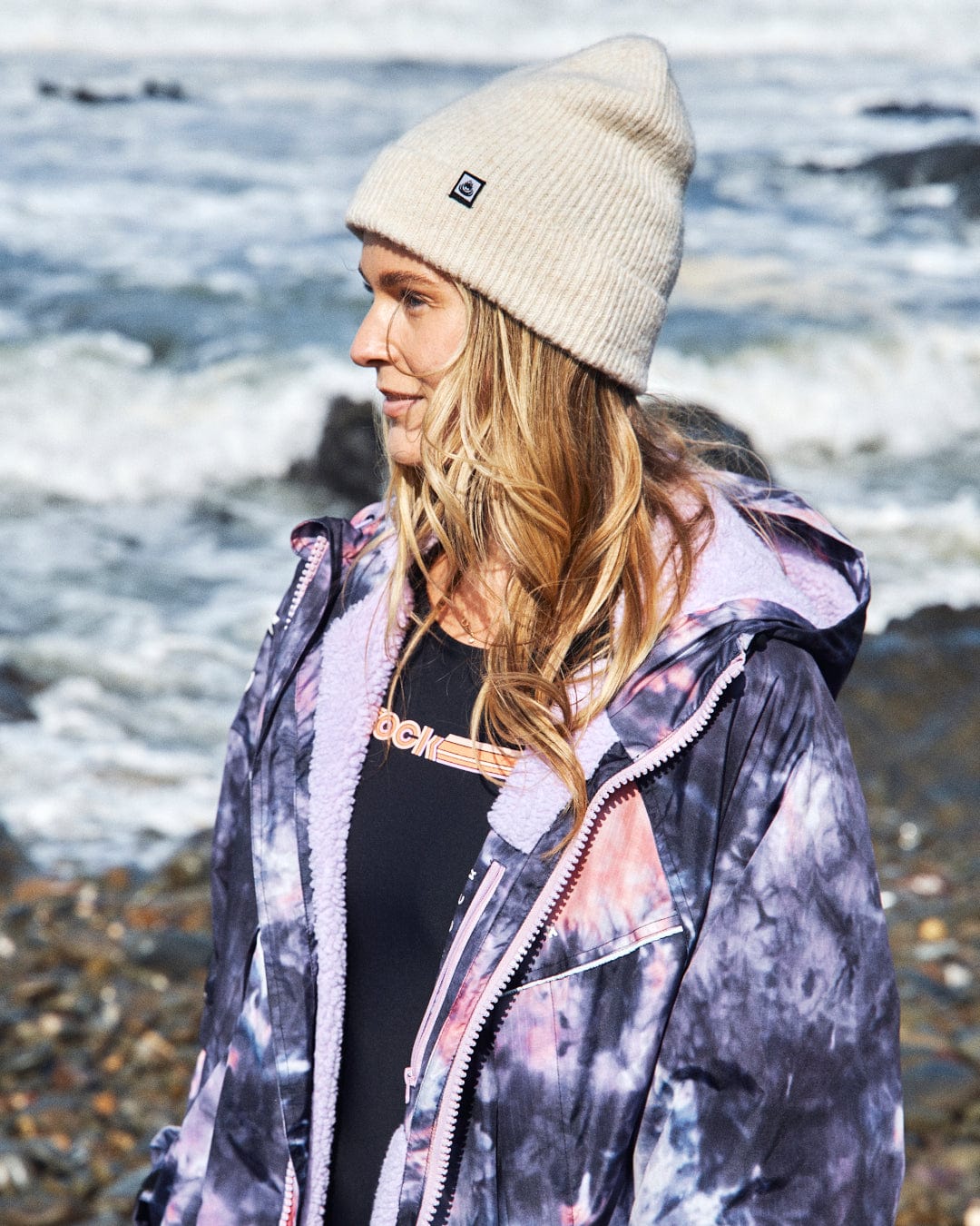 A person in a Saltrock Recycled Changing Robe in Pink Tie Dye and a beige beanie stands by the rocky shoreline, looking to the side.