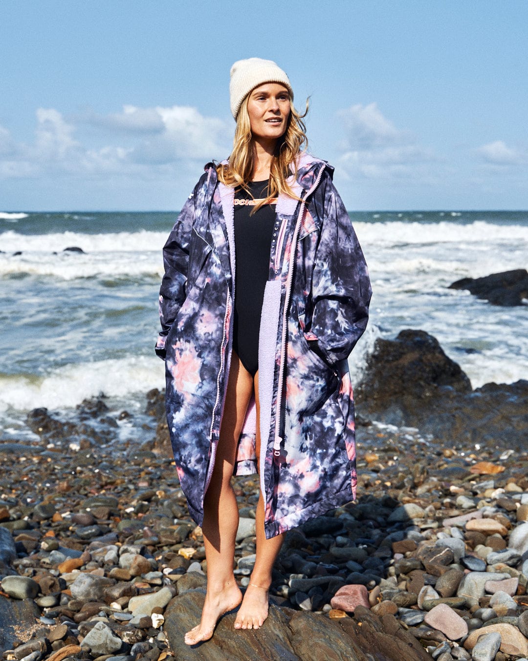 On a rocky beach, a person wears Saltrock's Recycled Changing Robe in Pink Tie Dye over a black swimsuit and sports a white beanie, with the ocean waves as the backdrop.