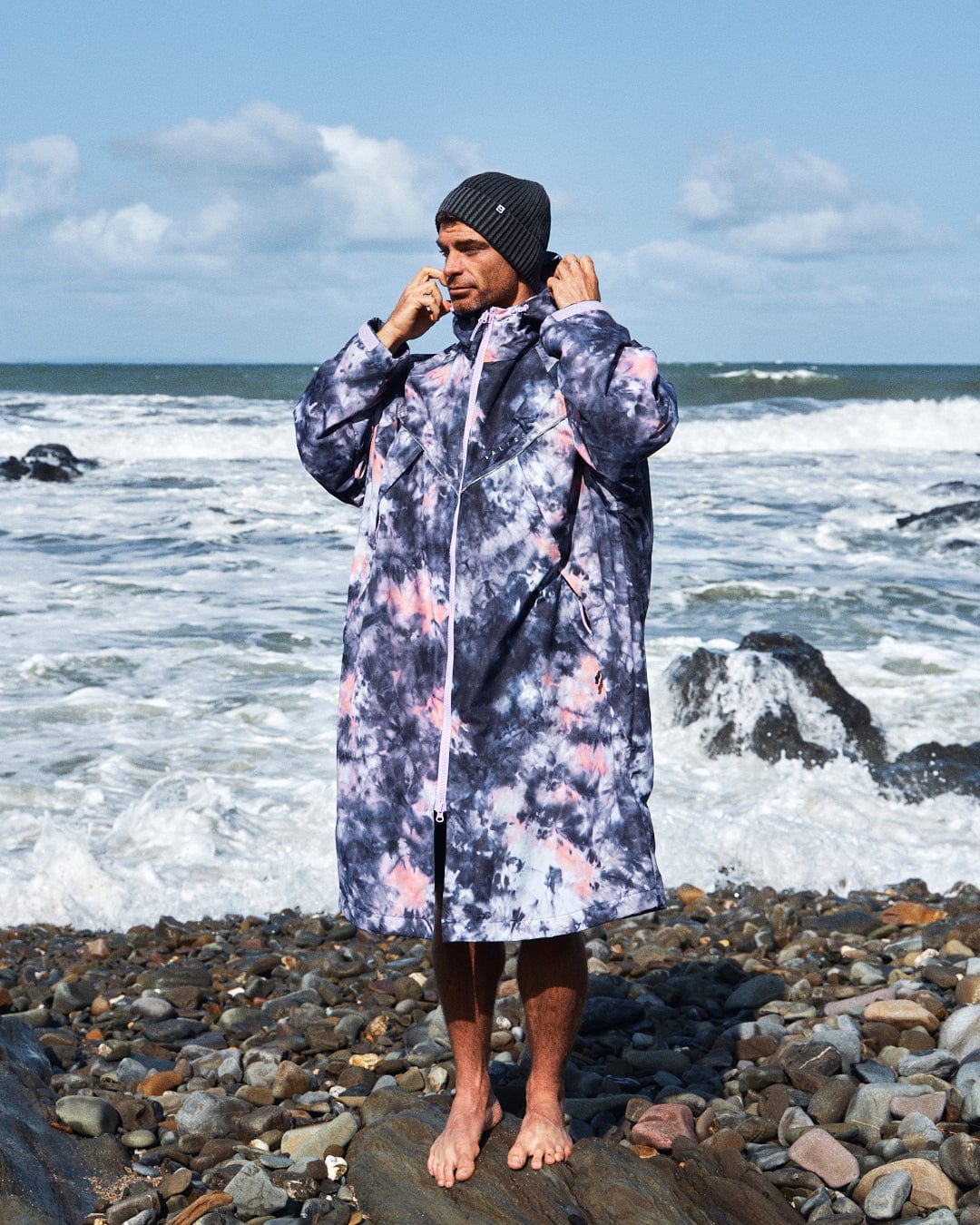 On a rocky beach with crashing waves, someone barefoot and wearing Saltrock's Pink Tie Dye Recycled Changing Robe and a beanie showcases their unique, style inspired by recycled materials.