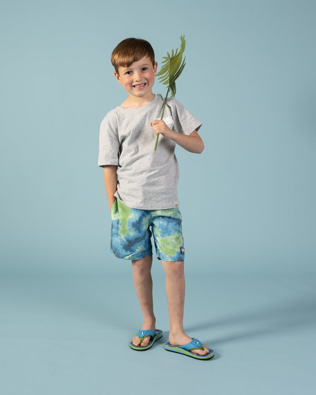 Young boy smiling on a light blue background, wearing a gray t-shirt and Saltrock Ocean Swim Kids Swim Shorts in blue with an elasticated waist, flip-flops, and holding a green palm leaf.