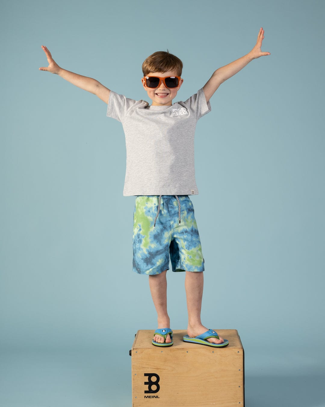A young boy in Ocean Swim - Kids Swim Shorts - Blue by Saltrock stands on a wooden box, arms outstretched, wearing sunglasses and a grey t-shirt against a plain blue background.