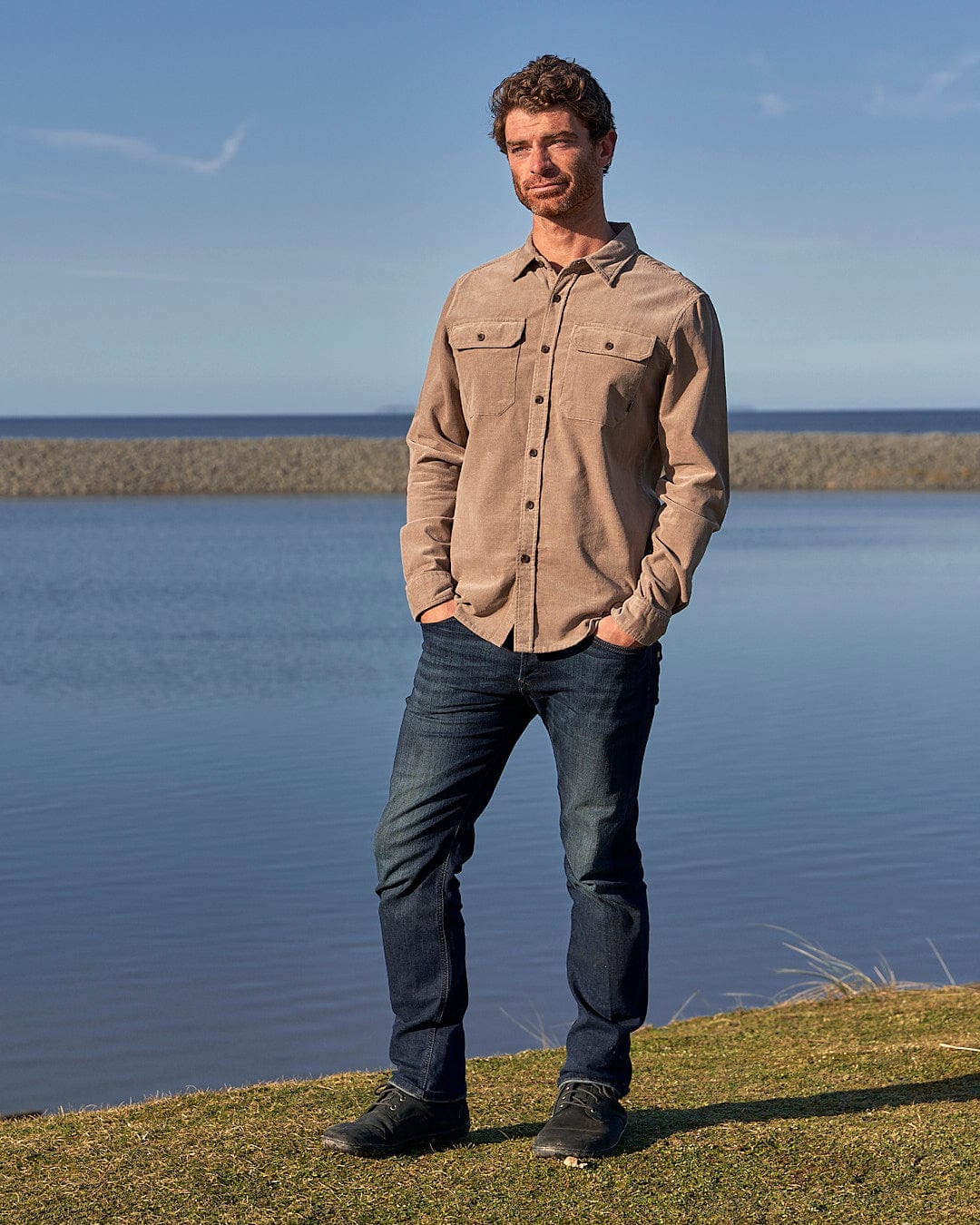 A man standing in front of a body of water wearing the Saltrock Ace - Men Corduroy Shirt in Light Brown.