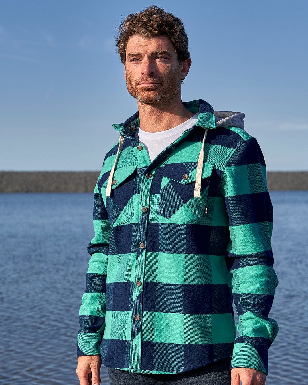 A man wearing the Saltrock Beale - Mens Heavy Weight Hooded Shirt - Dark Green standing by the water.