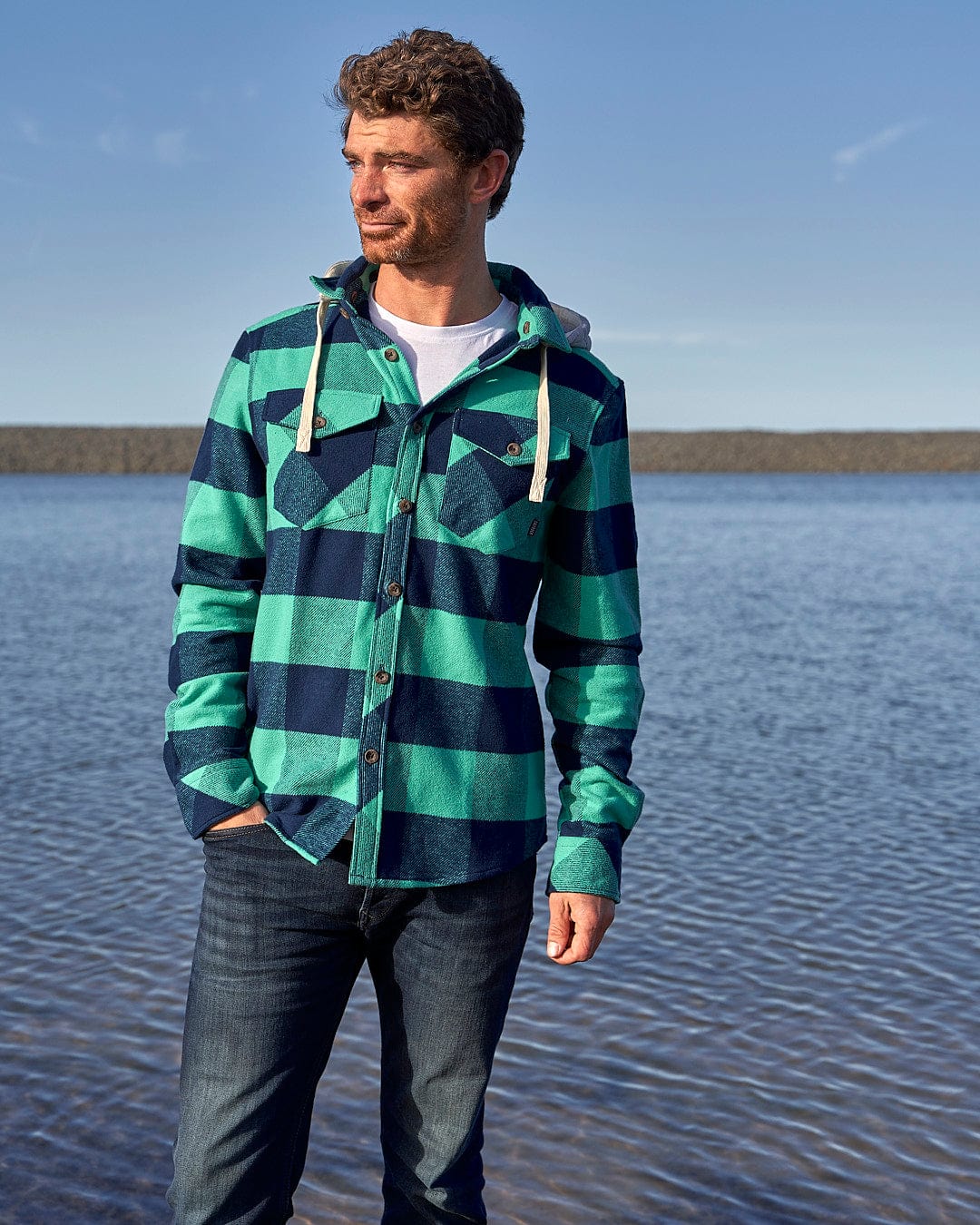 A man wearing a Saltrock Beale - Mens Heavy Weight Hooded Shirt - Dark Green standing by the water.