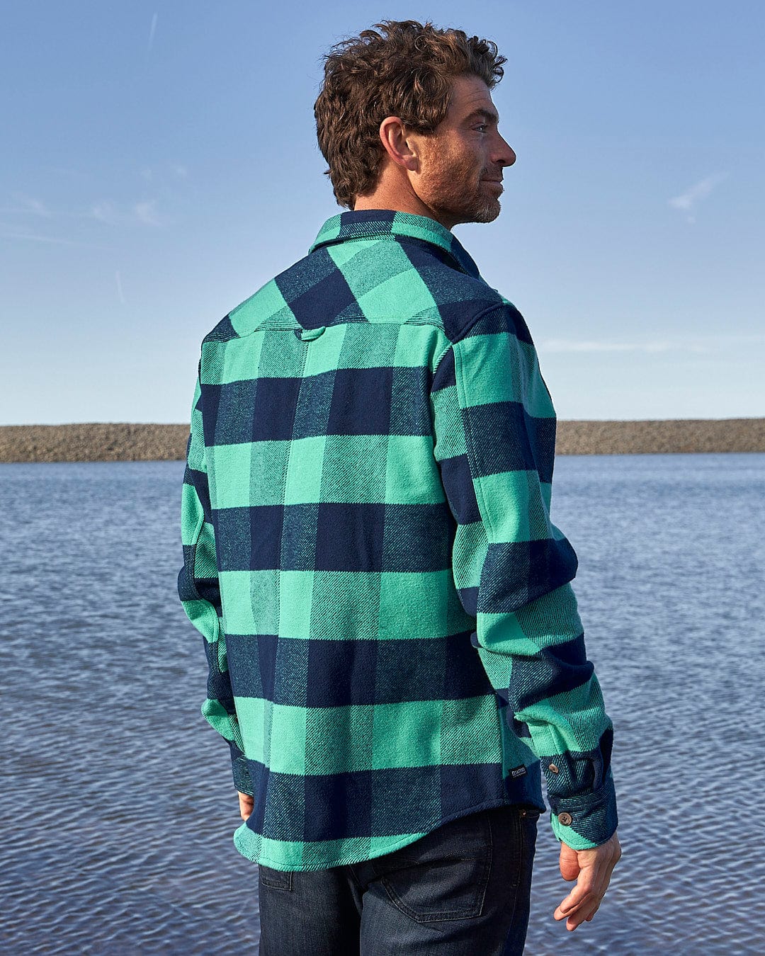 A man wearing a Saltrock Beale - Mens Heavy Weight Hooded Shirt - Dark Green standing by the water.