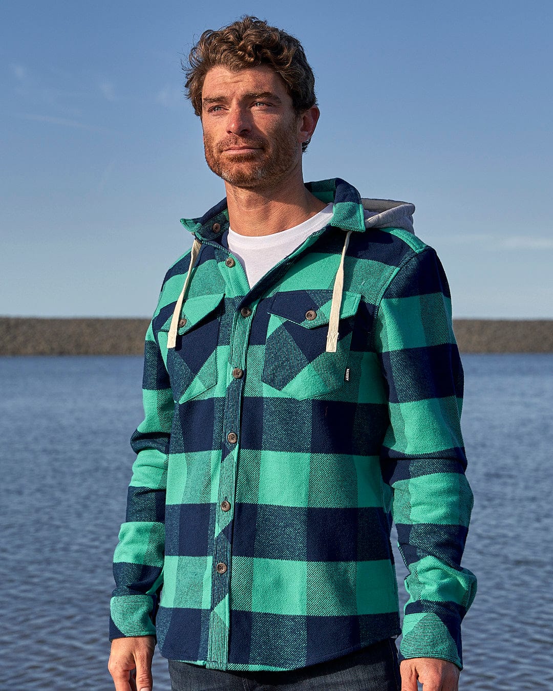 A man wearing a Saltrock Beale - Mens Heavy Weight Hooded Shirt - Dark Green flannel shirt standing by the water.