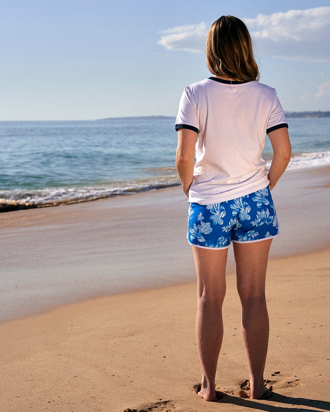A woman is standing on the beach wearing a Saltrock Celeste Stripe - Womens Ringer Tee - White, gazing at the ocean.