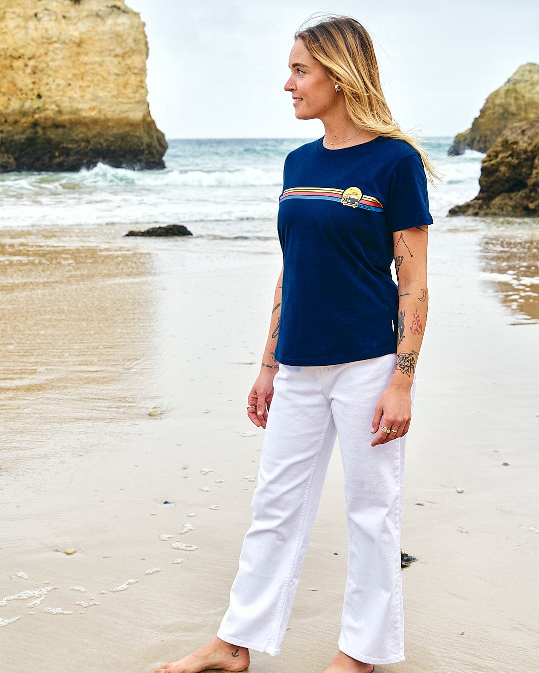 A woman standing on the beach wearing a Saltrock Celeste Stripe - Womens Short Sleeve T-Shirt - Blue and white pants.