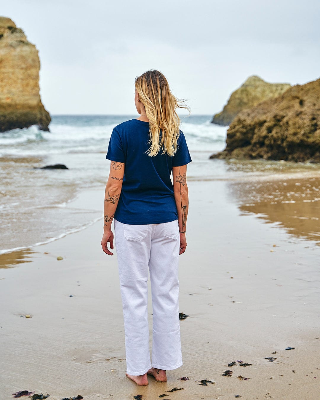A woman standing on a beach wearing white pants and a Saltrock Celeste Stripe - Womens Short Sleeve T-Shirt - Blue.