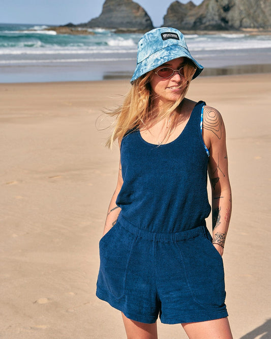 A woman wearing a Saltrock Gretchen - Womens Towelling Playsuit - Dark Blue and hat on the beach.