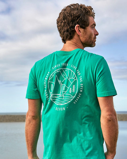A man wearing a Saltrock Devon Sailaway Outline - Mens Short Sleeve T-Shirt - Bright Green looking at the ocean.