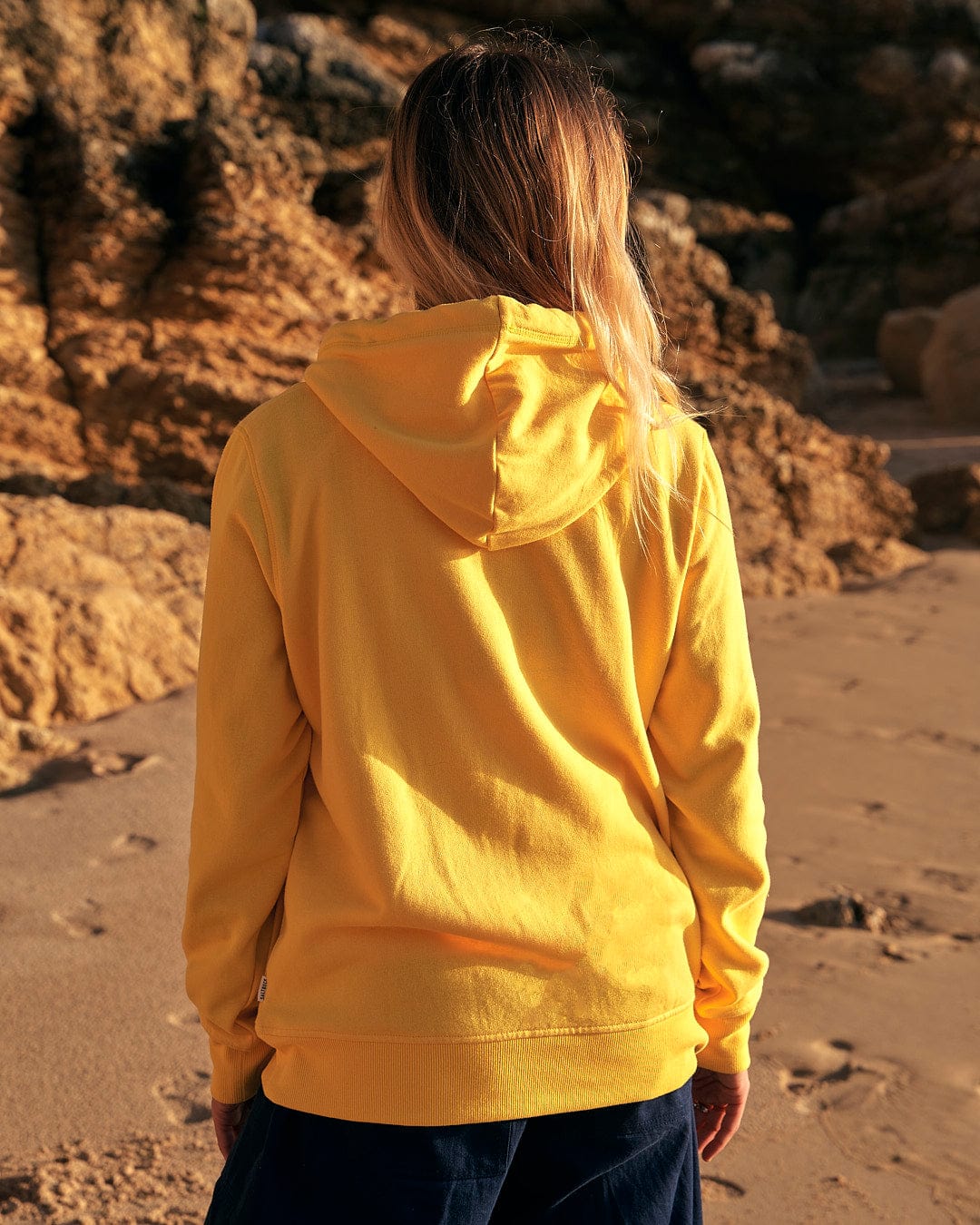 A woman wearing a Dreamseeker - Womens Pop Hoodie in yellow by Saltrock on the beach.