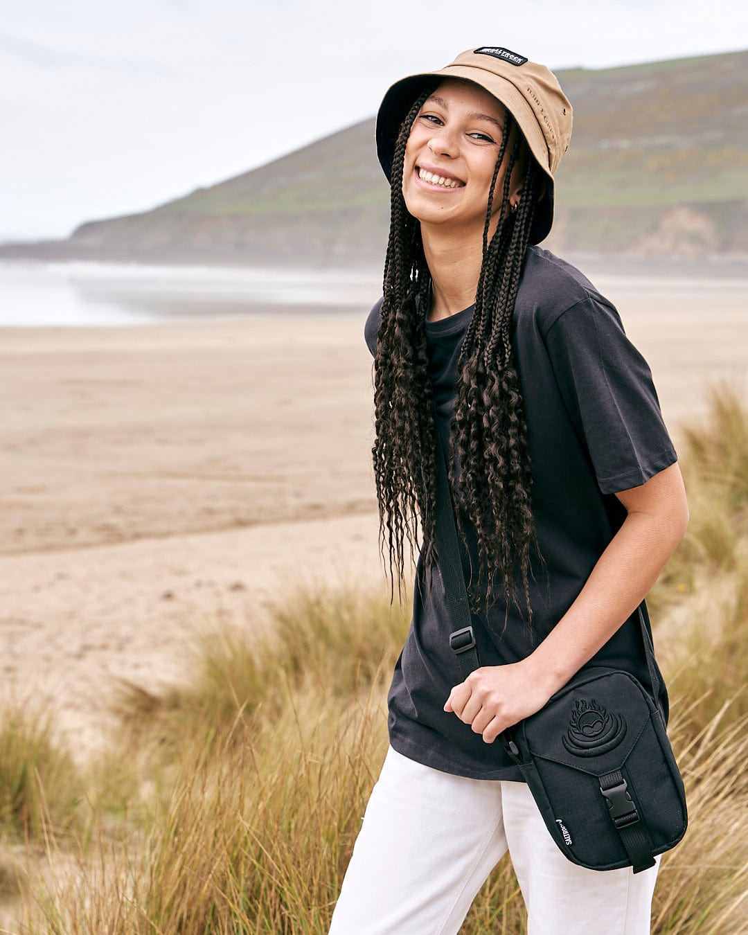 Young woman smiling on a beach with a hat and a Saltrock Festival Shoulder Bag in Black featuring adjustable strap.