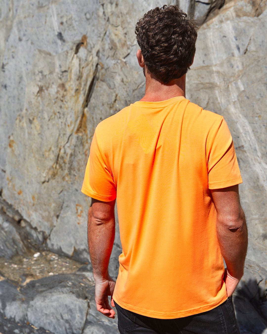 A man wearing a Saltrock Flame Tri - Mens Recycled Short Sleeve T-Shirt - Light Orange standing next to rocks.