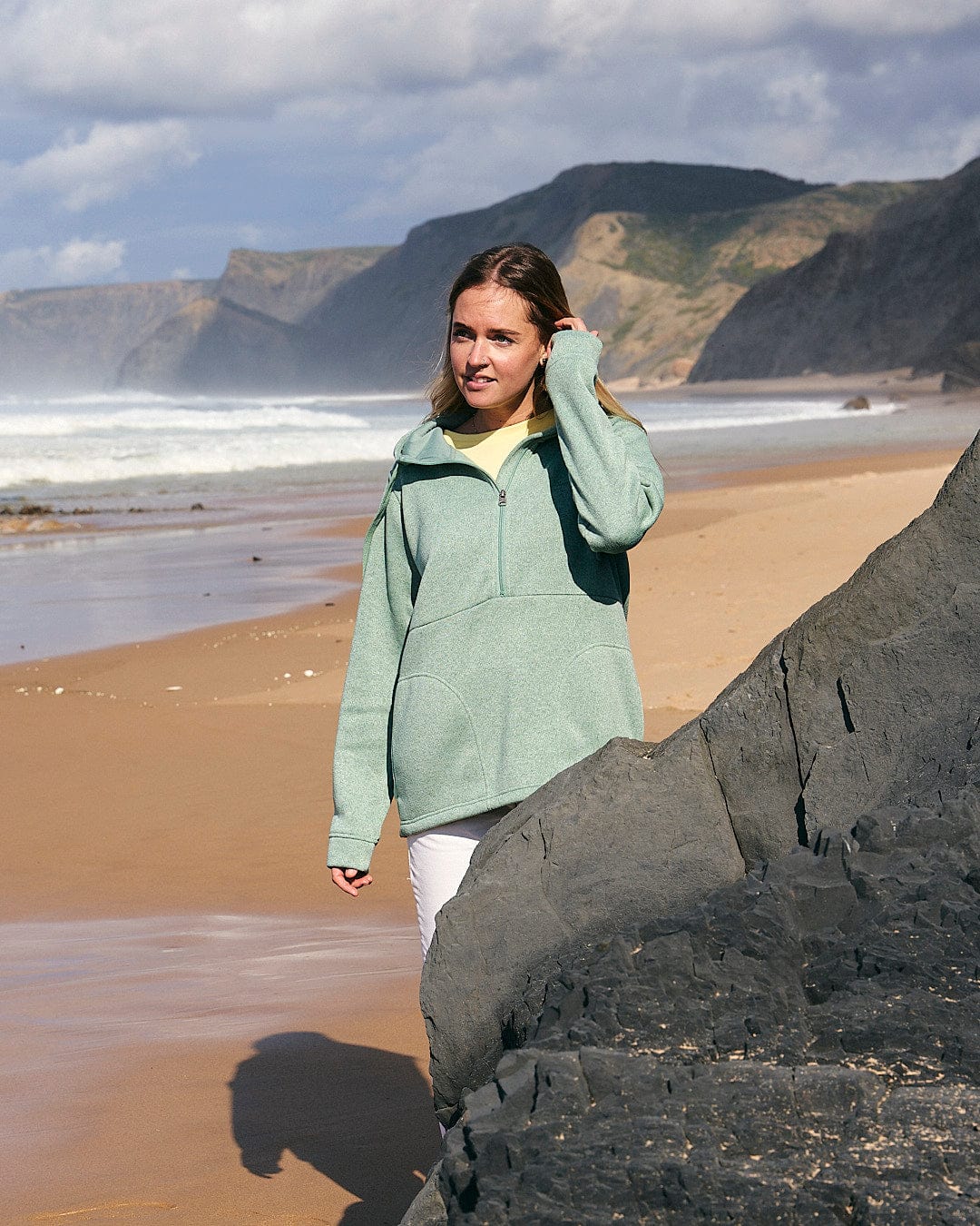 A woman standing on a beach next to a rock, wearing the Galaksea - Womens 1/4 Zip Fleece - Light Green by Saltrock.