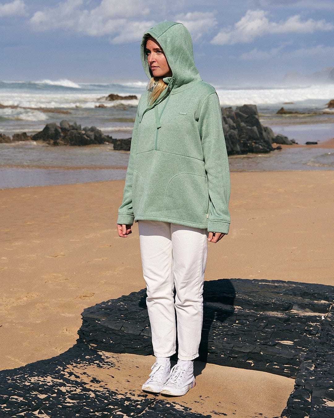 A woman wearing a Saltrock Galaksea - Womens 1/4 Zip Fleece - Light Green hoodie standing on a beach.