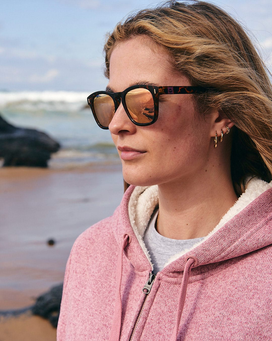 A woman wearing sunglasses and a pink hoodie, finding comfort in her Saltrock Galak - Womens Fur Lined Hoody - Pink, enjoying an adventure on the beach.