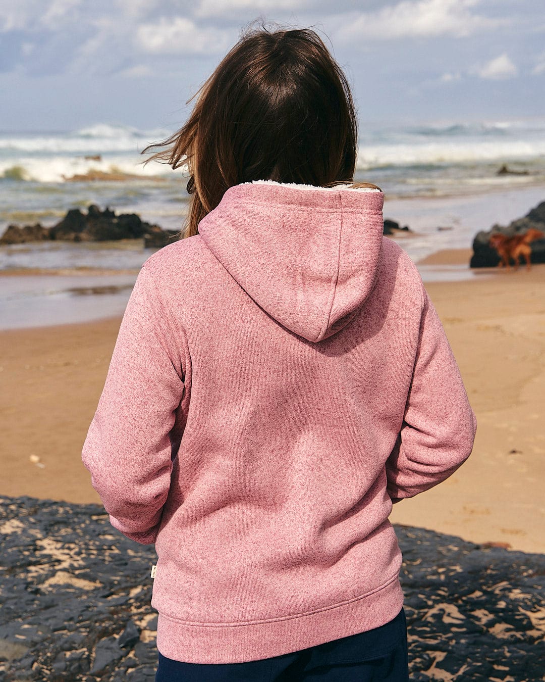 A woman wearing the Galak - Womens Fur Lined Hoody - Mid Pink by Saltrock standing on the beach.