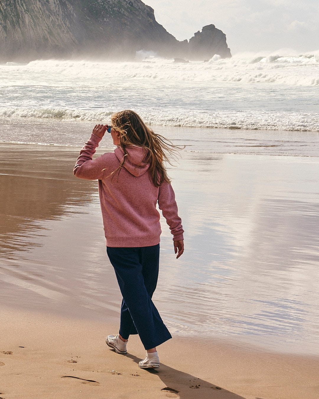 A woman wearing a Saltrock Galak - Womens Fur Lined Hoody - Pink walks on a beach near the ocean, finding both comfort and adventure.