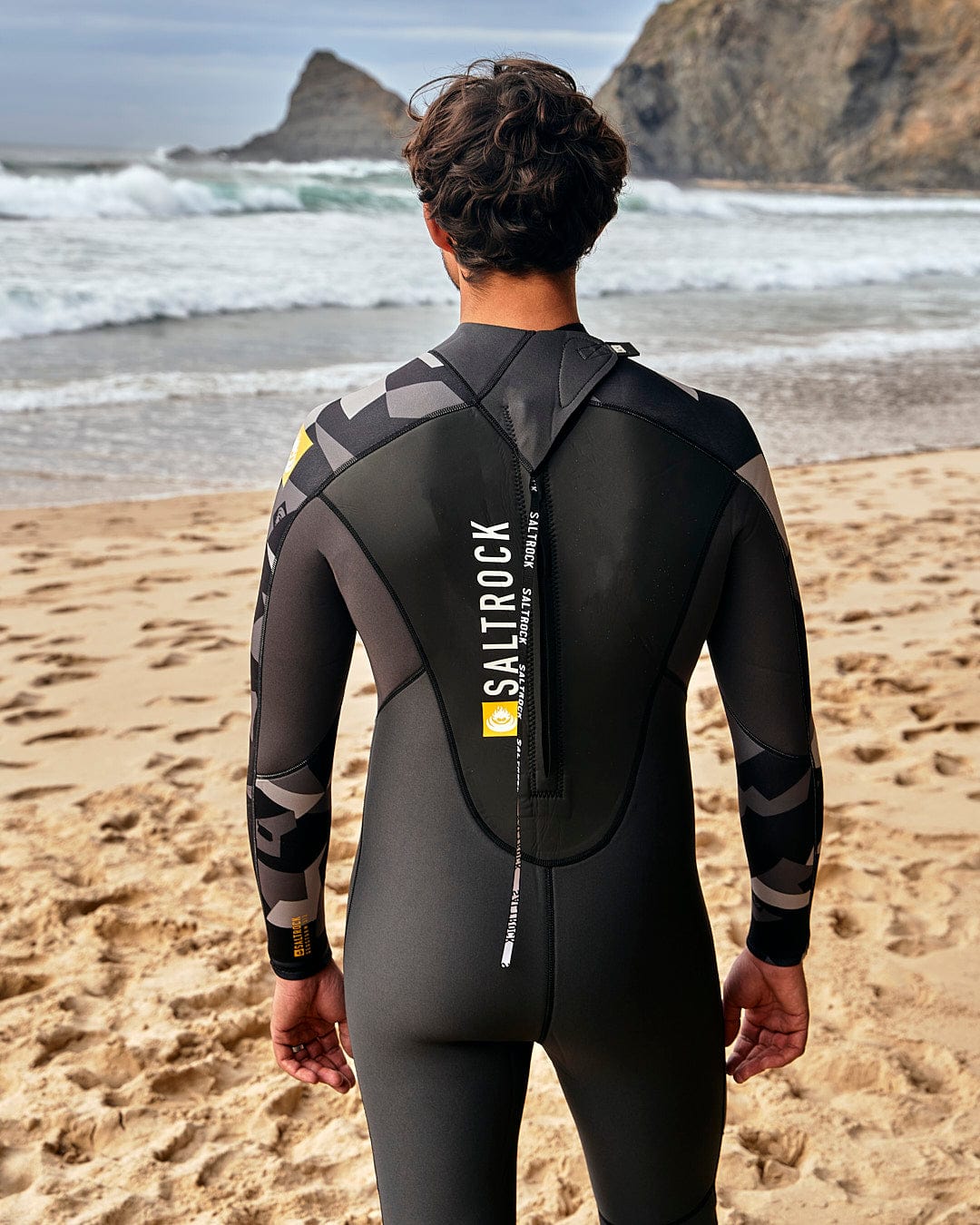 Back view of a man wearing a Saltrock Geo Camo - Mens 3/2 Full Wetsuit - Black on the beach.