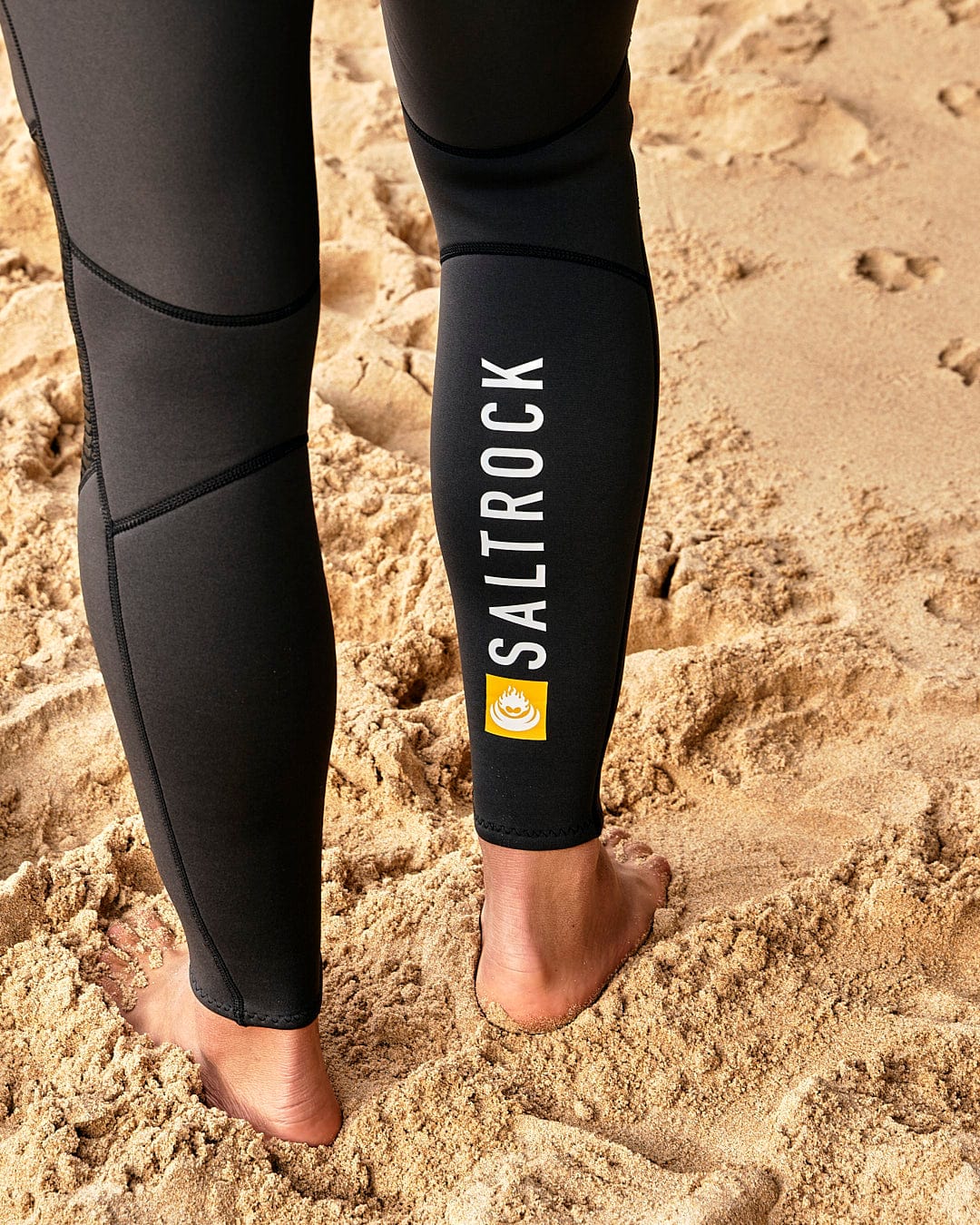 A person wearing a Saltrock Geo Camo - Mens 3/2 Full Wetsuit - Black standing on the sand.