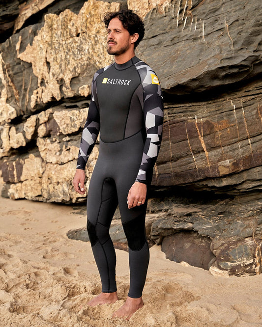 A man is standing on the beach wearing a Saltrock Geo Camo - Mens 3/2 Full Wetsuit - Black.