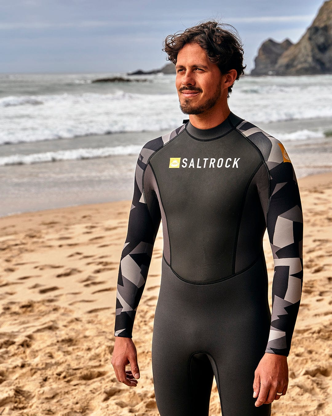 A man standing on the beach wearing a Saltrock Geo Camo - Mens 3/2 Full Wetsuit - Black.