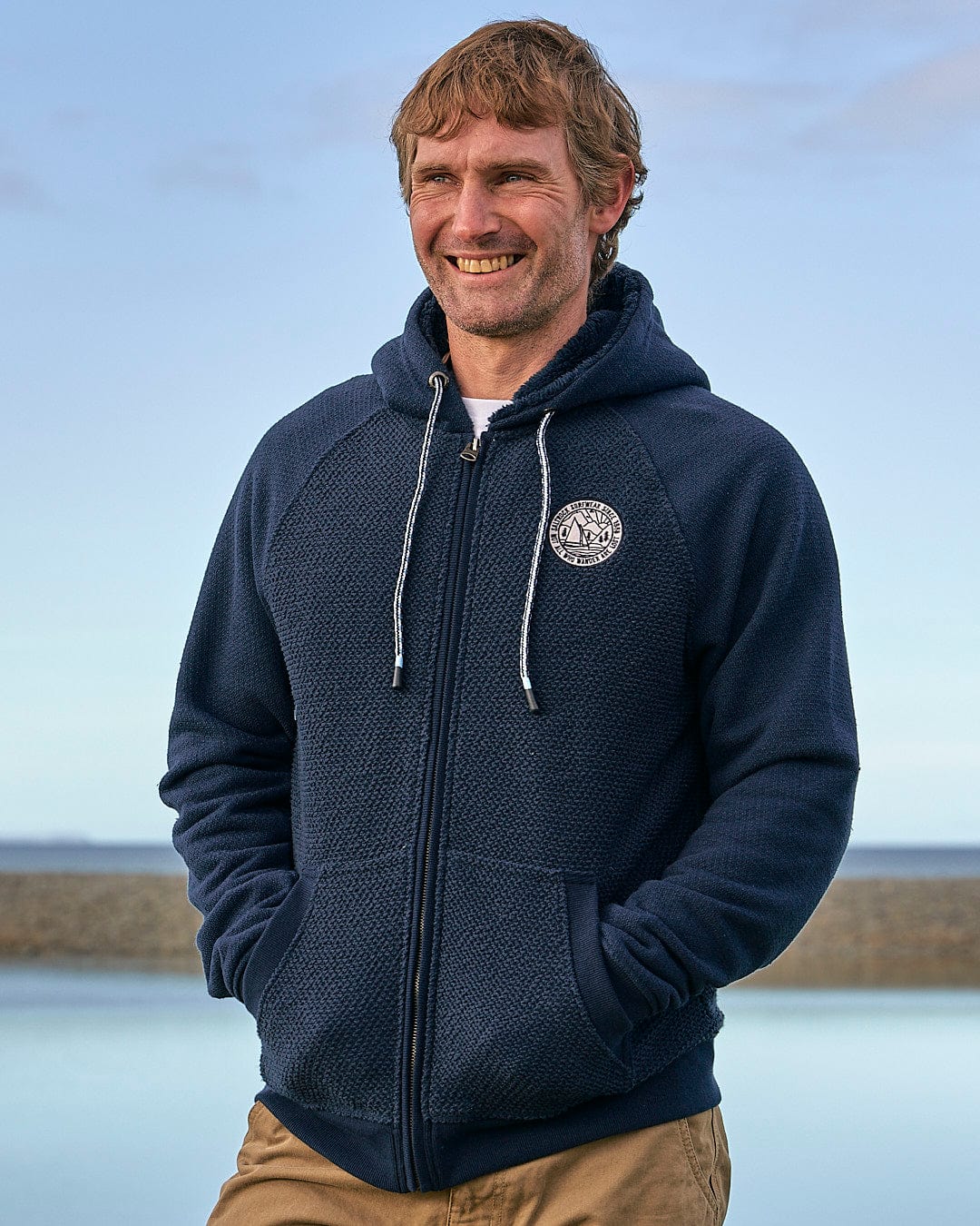 A man is smiling in front of a body of water while wearing the Saltrock Hall - Mens Orkney Borg Lined Hoodie in Dark Blue.